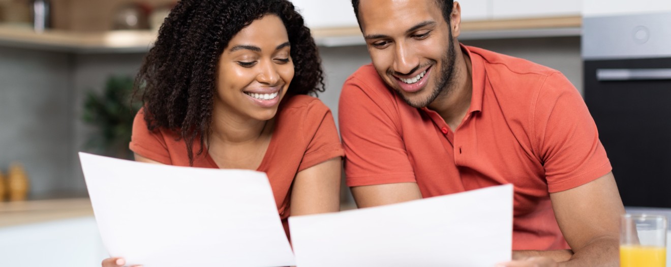Junges verheiratetes Paar sitzt lächelnd am Tisch und prüft gemeinsam Unterlagen.