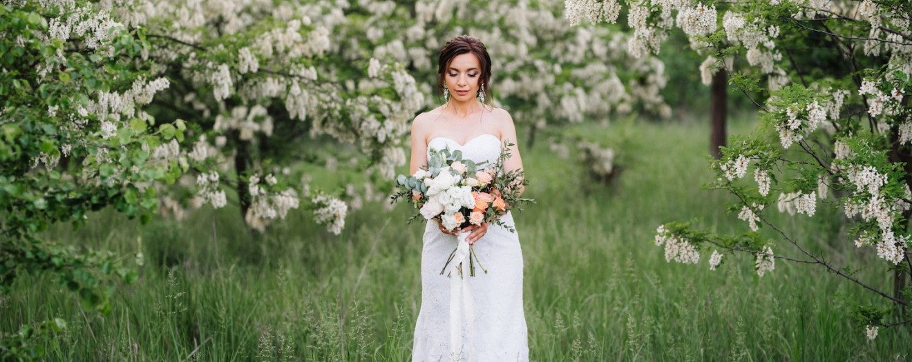 Braut in weißem Kleid steht inmitten einer blühenden Frühlingslandschaft und hält einen eleganten Brautstrauß aus weißen und apricotfarbenen Blumen.
