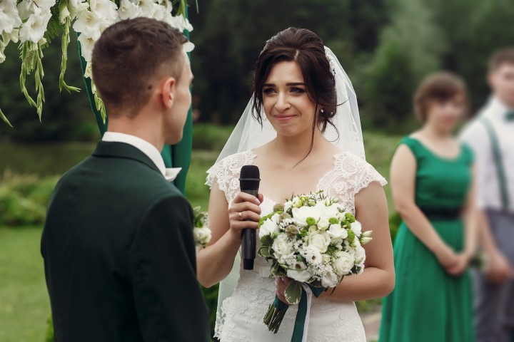 Emotional gerührte Braut im weißen Kleid hält einen Blumenstrauß und ein Mikrofon und spricht mit Tränen in den Augen zu ihrem Bräutigam während der Trauung im Freien.