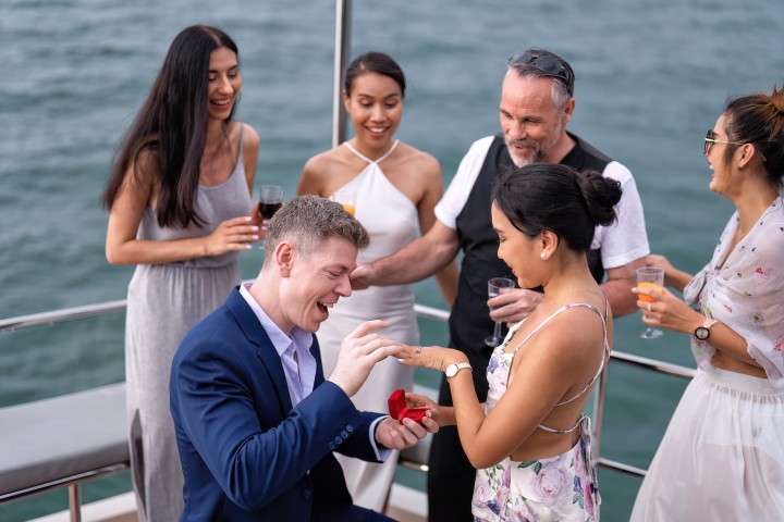 Ein Mann kniet bei einem Heiratsantrag auf einem Boot und steckt seiner Partnerin feierlich den Verlobungsring an. Um sie herum jubeln Freund:innen mit Sekt- und Weingläsern in der Hand.