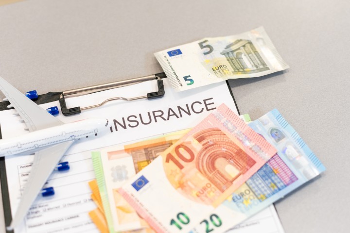 Versicherungspapier mit der Aufschrift ‚Insurance‘, darauf Euro-Geldscheine und ein Modellflugzeug – Symbolbild für Versicherungsoptimierung.