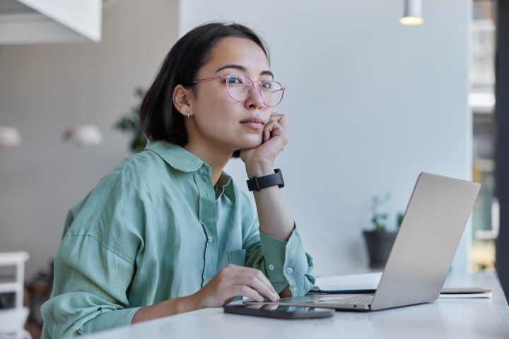 Eine Frau sitzt mit nachdenklichem Blick vor Laptop und Smartphone