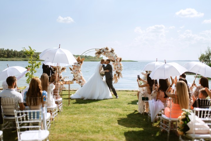 Ein Brautpaar küsst sich bei einer freien Trauung am See, umgeben von blühendem Blumenschmuck und sommerlich gekleideten Gästen mit Sonnenschirmen – ein stimmungsvolles Beispiel für eine Hochzeit im Sommer und ihre Besonderheiten im Jahreszeitenvergleich.